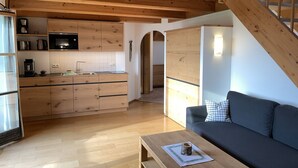 Una cocina moderna con armarios de madera, un microondas integrado y una cafetera. Hay una mesa de comedor de madera con una taza encima, y un sofá de color oscuro en la zona de estar.