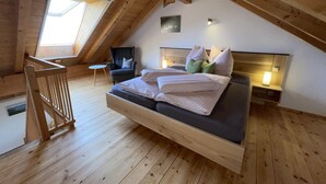 Un dormitorio tipo loft con techo inclinado, cama con ropa de cama estampada, una silla, mesita de noche con lámpara y tragaluz.