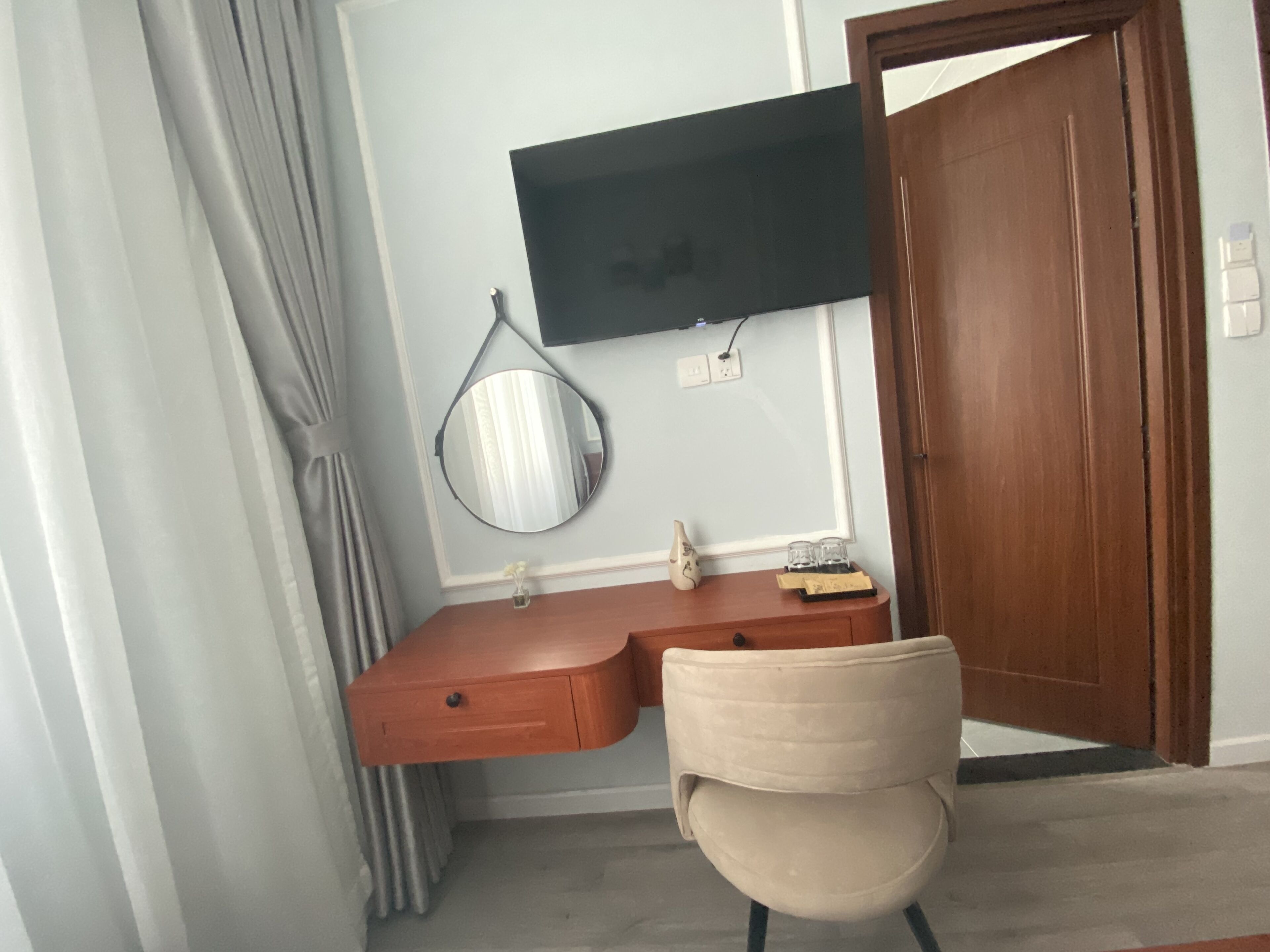 Une pièce comprenant un bureau en bois, une chaise, un téléviseur à écran plat fixé au mur, une porte en bois et une fenêtre avec des rideaux.