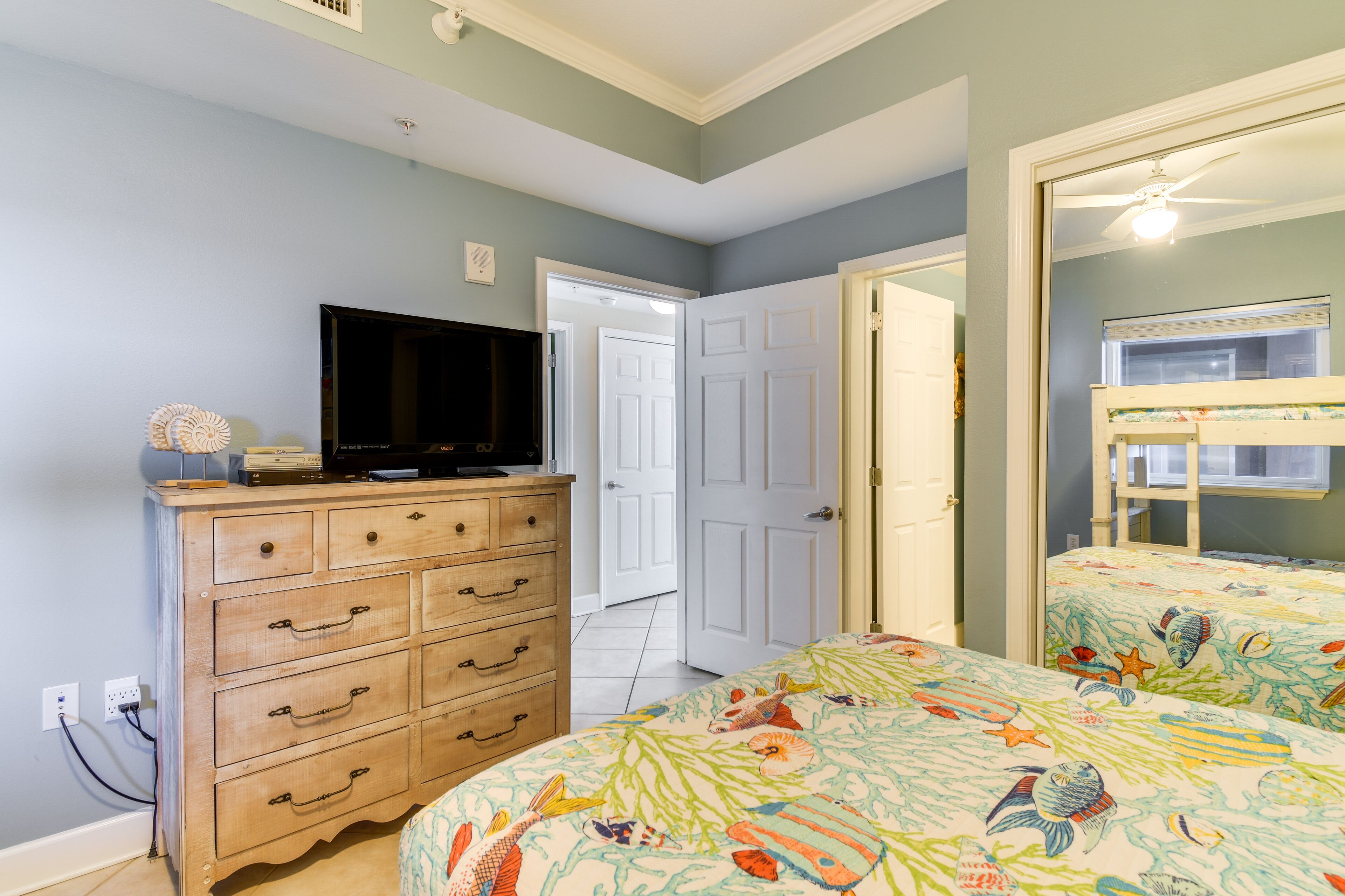 A bedroom with a wooden dresser, a television, a bunk bed with a fish-themed bedspread, and a door leading to another room.