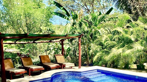Una piscina con borde de azulejos, rodeada por una pérgola de madera y sillones de descanso.