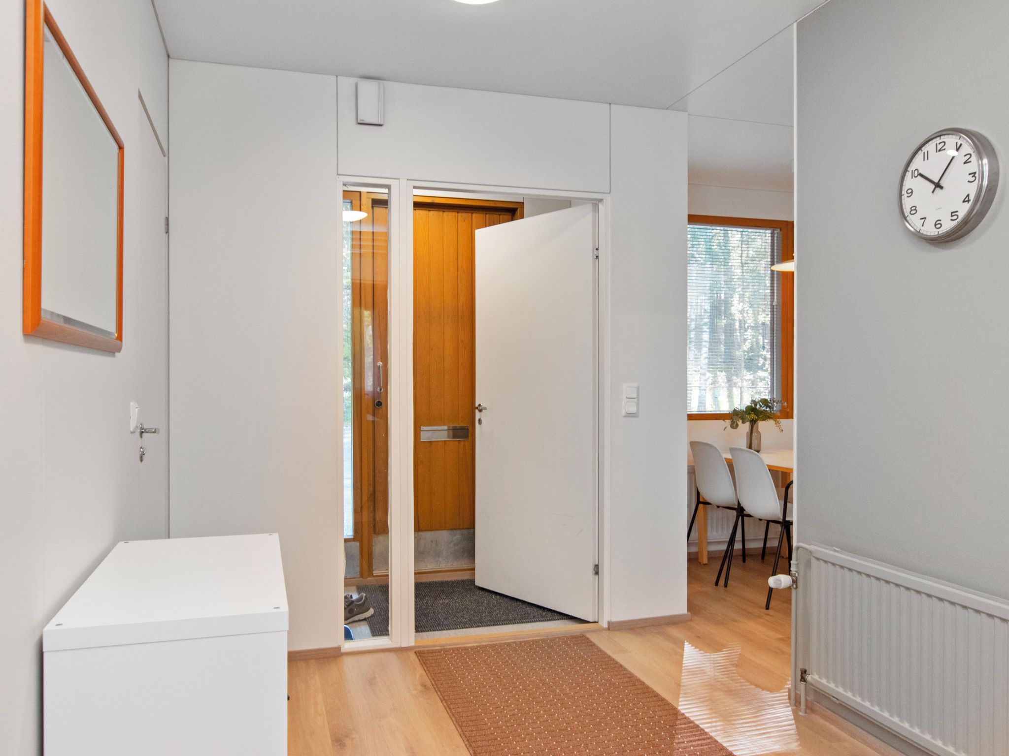 A hallway with a white door leading to a room with a wooden door, a dining area with chairs, and a wall clock.