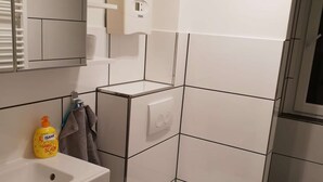Ett modernt badrum med en vit tvättställ, en toalett och en bidé. Det finns en handduksställning med ett handduk och en flaska tvål. Väggarna är kaklade i vitt och grått, och det står en liten papperskorg bredvid toaletten.