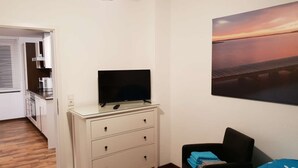 Ett hotellrum med en säng, en tv på en byrå, en tavla som föreställer en solnedgång och ett litet kök.