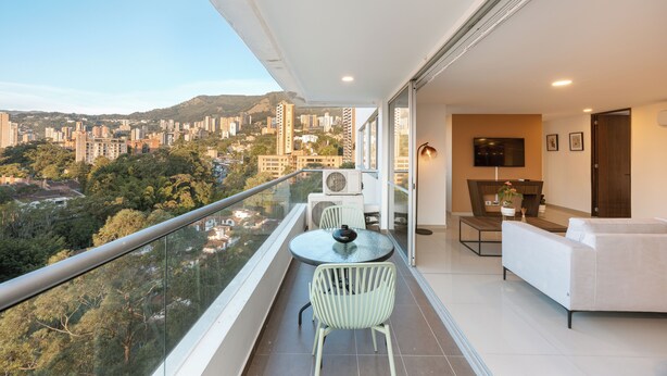 A balcony with a glass railing, a dining table with chairs, a sofa, and a TV mounted on the wall.