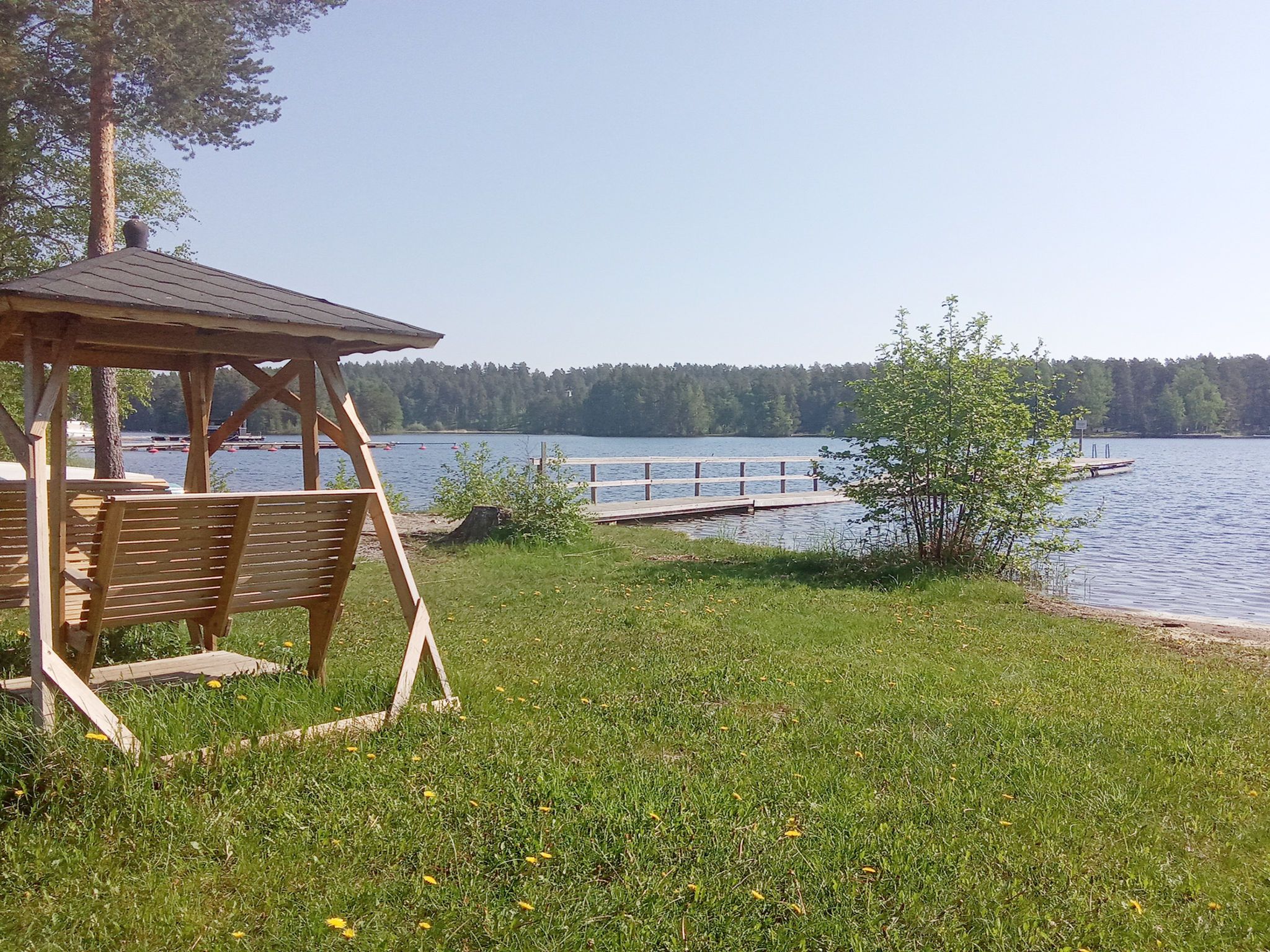 A wooden gazebo with benches, a dock extending into a lake, and a forested shoreline.