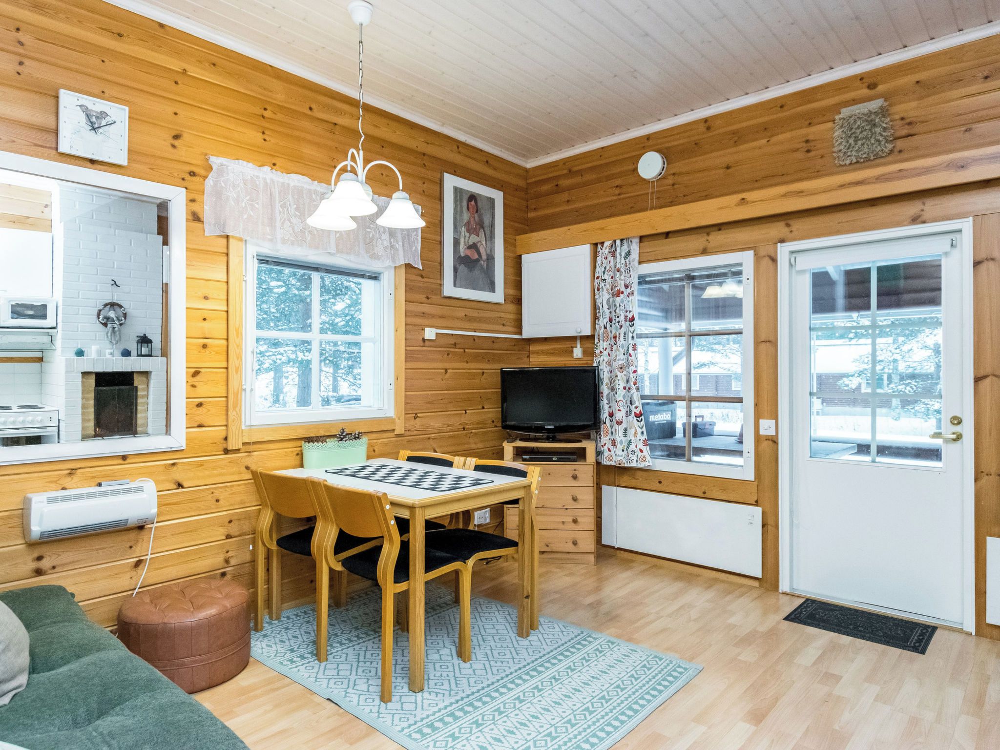 A cozy cabin interior with wooden walls, a fireplace, a dining table with chairs, a television, and a large window.