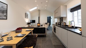 A modern kitchen with a central island, a dining area with a table set for two, and a cozy seating area with a fireplace. - The Bothy (Ulverston)