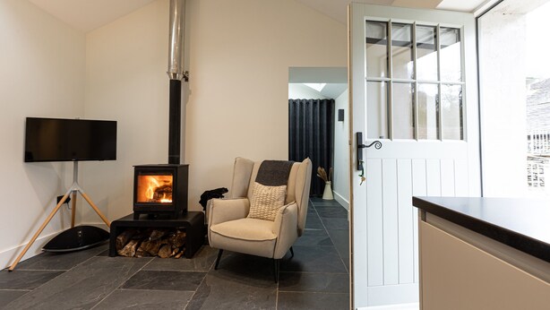 A cozy living room with a fireplace, a comfortable armchair, a flat-screen TV, and a door leading to another room. - The Bothy (Ulverston)