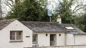 A white building with a sloped roof, a chimney emitting smoke, a small window, and a door. - The Bothy (Ulverston)