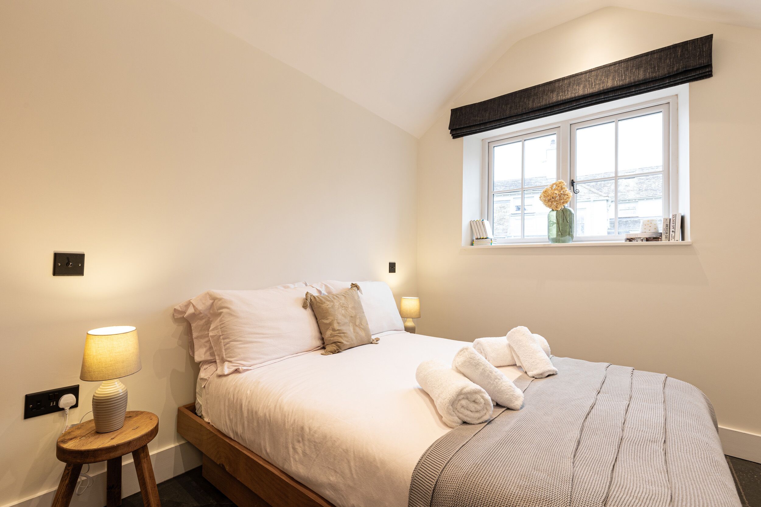 Een net opgemaakt bed met witte lakens en een grijze plaid, een bedlampje en een raam met uitzicht.