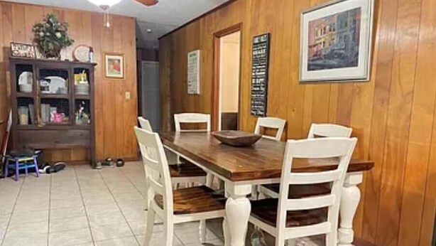 Un comedor con una mesa de madera y sillas blancas, un mueble de madera con puertas de cristal y un ventilador de techo.