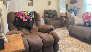 Una sala de estar con un sillón marrón, un sofá y una mesita. Hay dos cuadros en la pared.