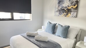 Un lit bien fait, avec une couverture grise et deux serviettes blanches, un oreiller gris et un oreiller bleu, le tout posé sur une tête de lit blanche.