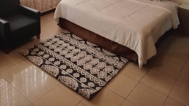 A bedroom with a bed, a chair, a patterned rug, and curtains. - Fresh Air Lodge (Accra)