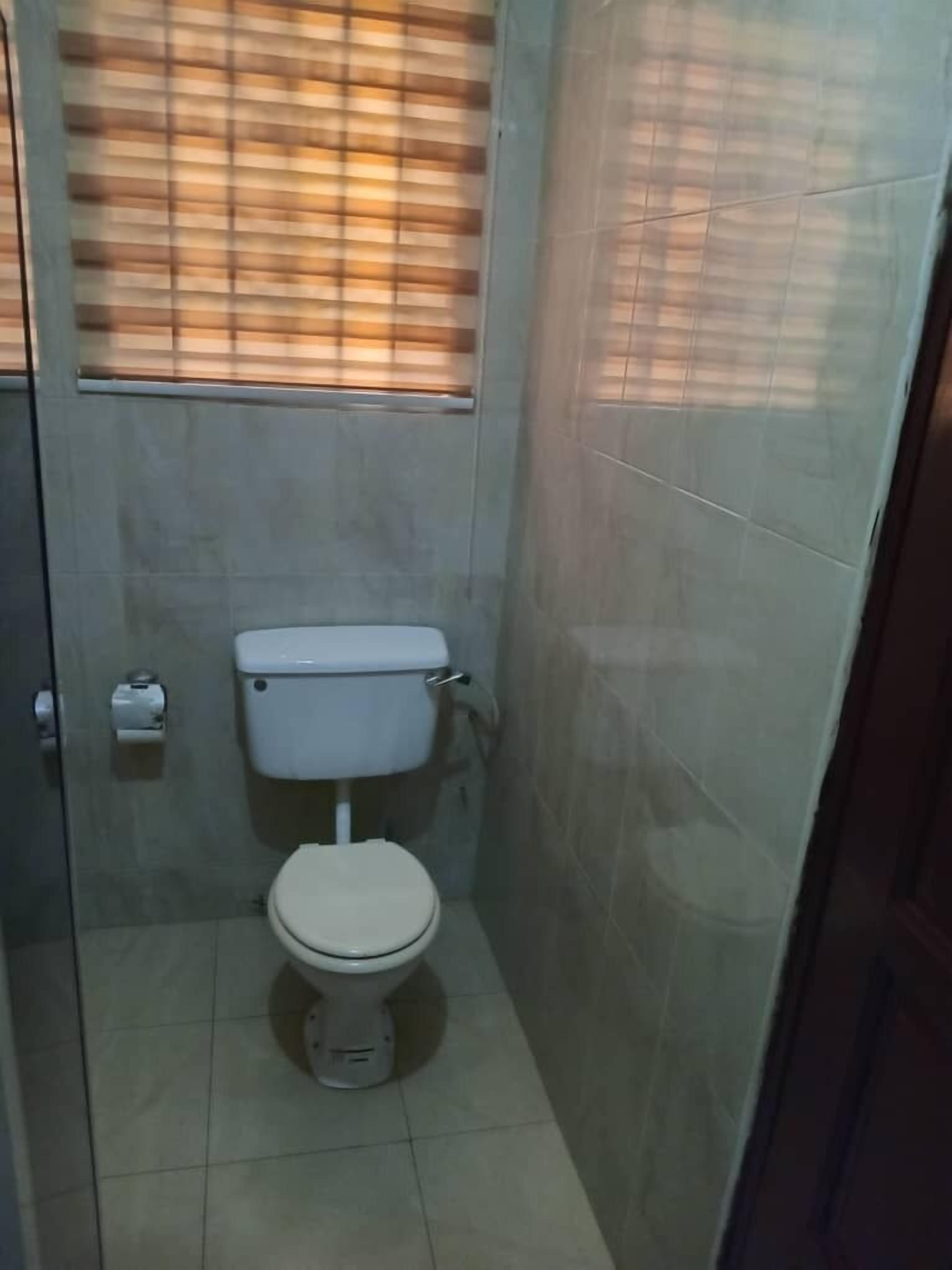 Un baño pequeño con azulejos, un inodoro, un rollo de papel higiénico y una ventana con persianas.