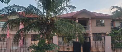 A two-story house with a gated entrance, a palm tree in the driveway, and a clear sky.