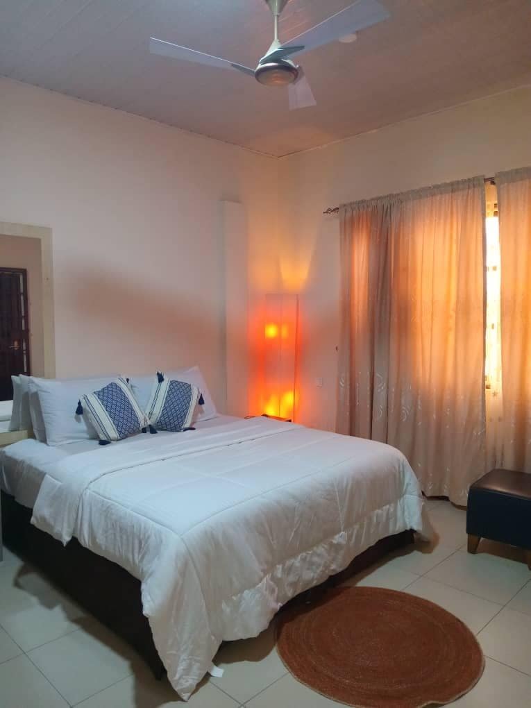 Una habitación de hotel con una cama, un ventilador de techo, una mesita de noche y una ventana con cortinas.