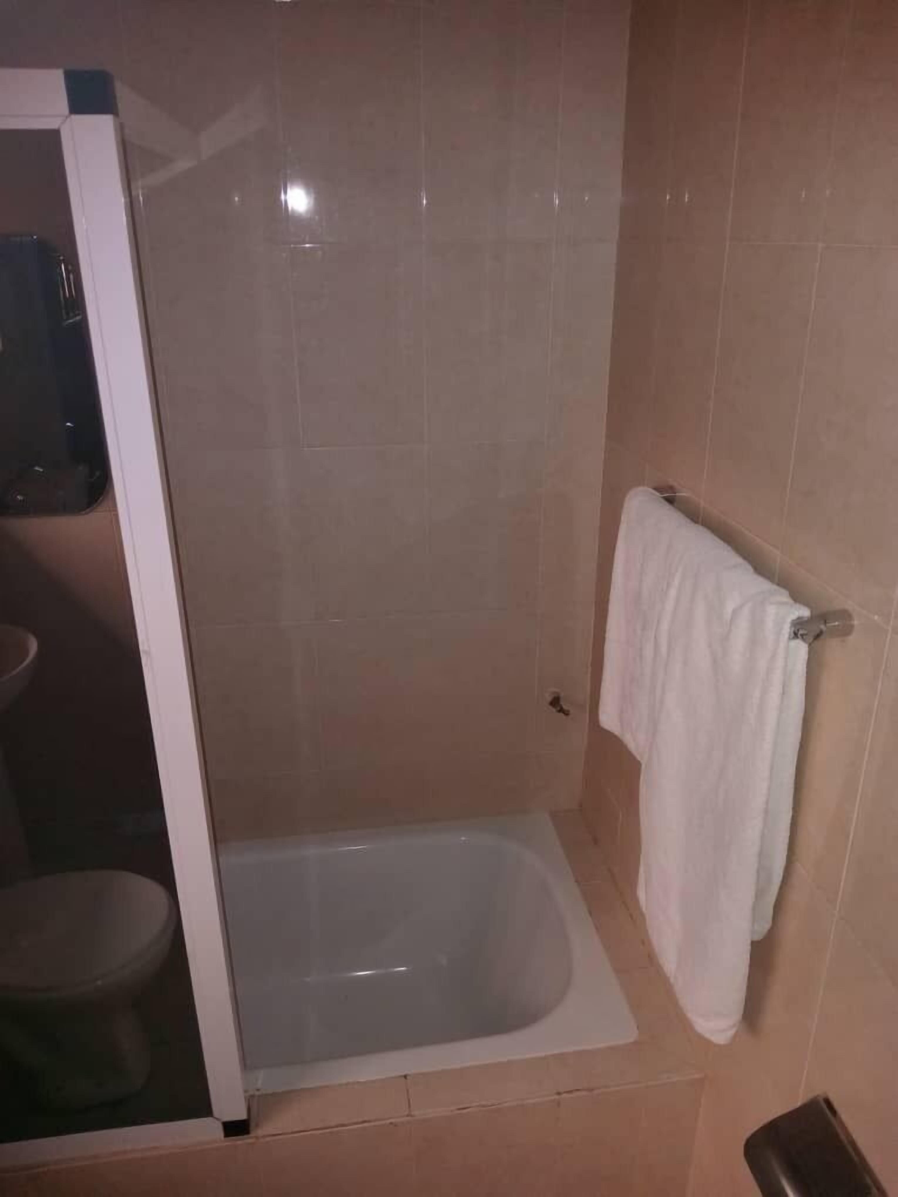 A small, enclosed shower area with a bathtub, a white towel hanging on a rack, and a toilet visible through a glass partition.