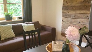 Ein modernes Wohnzimmer mit einem braunen Sofa, einem Esstisch mit einer Holzschale und einem Blumenstrauß, sowie einem großen Fenster mit Vorhängen.