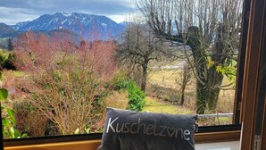 Ein Fenster mit einem Kissen mit dem Aufdruck „Kuschelzone“ und Blick auf Berge und Bäume.