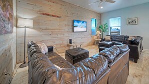 A modern living room with a wooden wall, a ceiling fan, a flat-screen TV, and a large leather sofa. - 6BDR, Balcony w/ Bay View, Shared Pool + Boat Dock | Keeper Red by AvantStay (Corpus Christi)