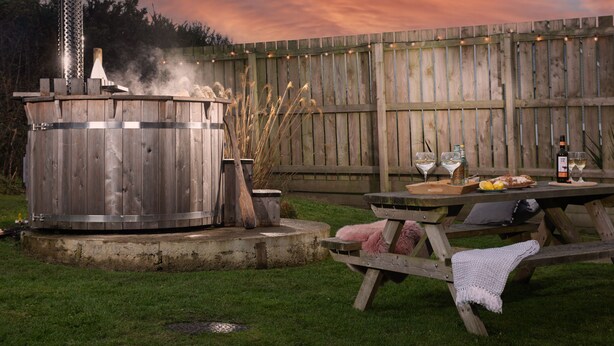 A wooden outdoor grill with a chimney, a picnic table with food and drinks, and a wooden fence in the background. - Wheal Prosper Lodge (Helston)