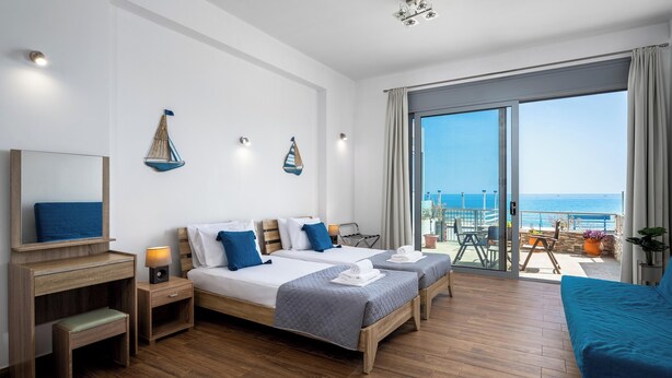 A hotel room with two beds, a desk, a chair, a balcony with a view of the sea, and a large window.