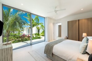 Una camera da letto moderna con un'ampia porta scorrevole in vetro che si apre su una vista di un giardino tropicale, una ventola a soffitto e un letto con molti cuscini.