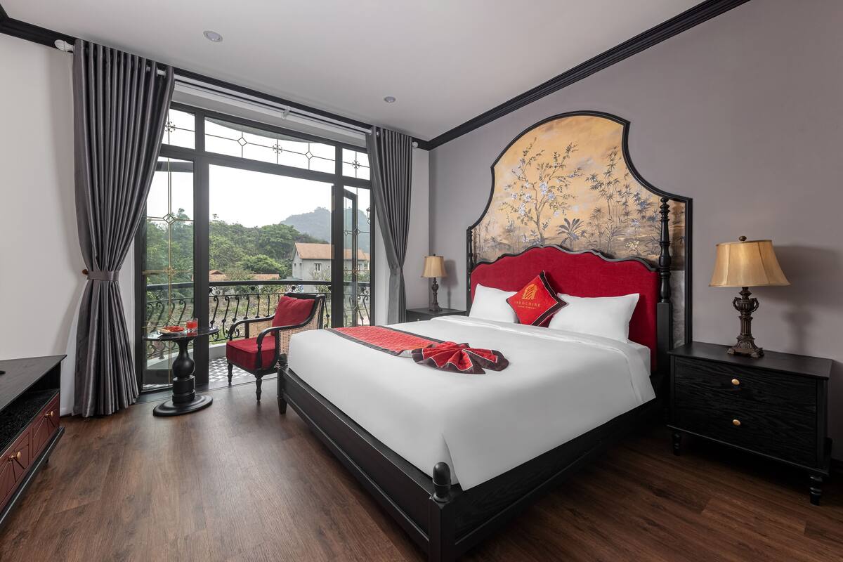 A hotel room with a large bed, a balcony with a table and chairs, and a view of greenery outside.