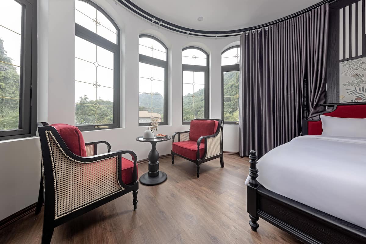 A hotel room with a large bed, two chairs, a small table, and large windows offering a view of greenery.