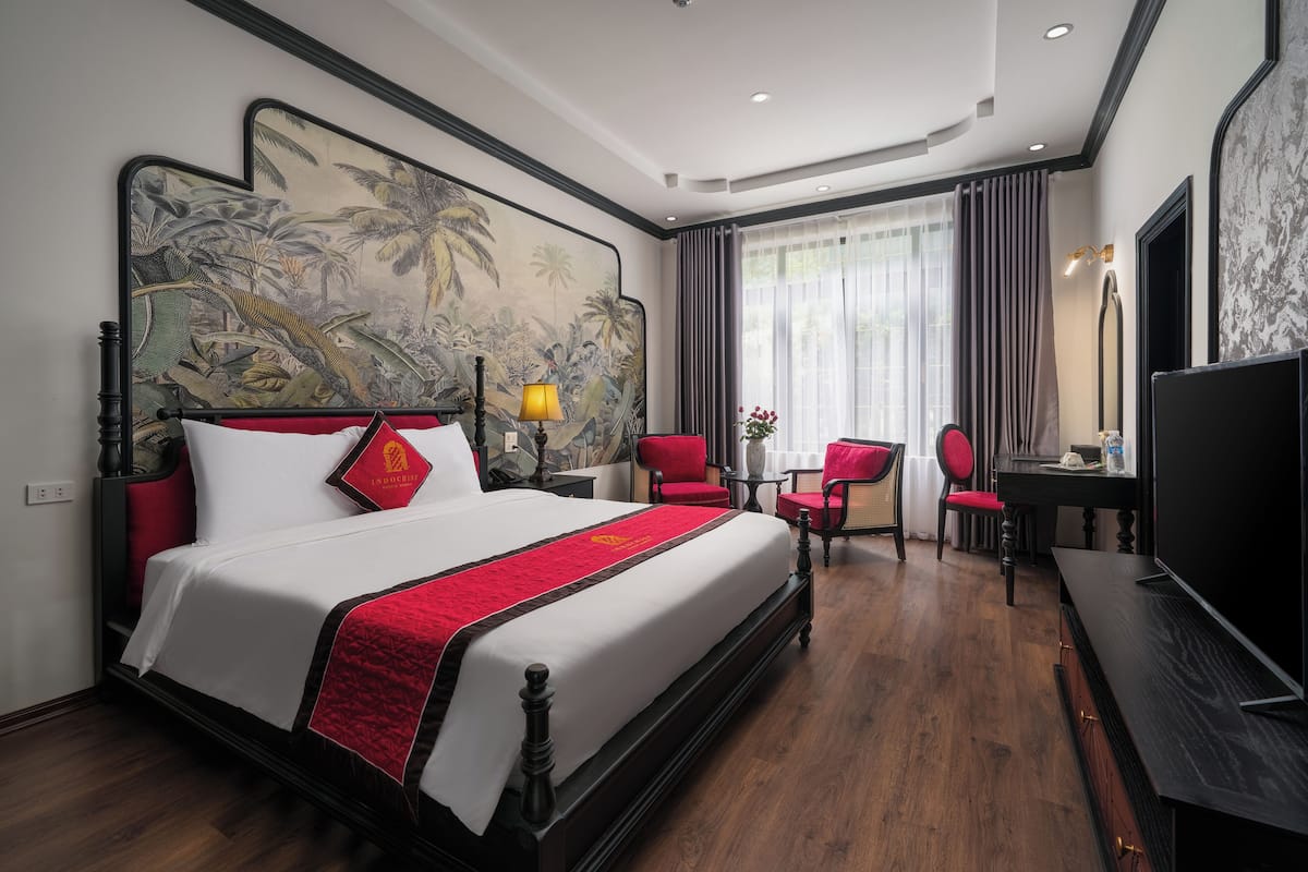 A hotel room with a large bed, a desk, two chairs, a TV, and a painting on the wall.