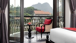 A balcony with a red chair, a small table with a drink, and a view of a mountain and a house. - Indochine Boutique Hotel Ninh Binh (Nam Hoa Lu)