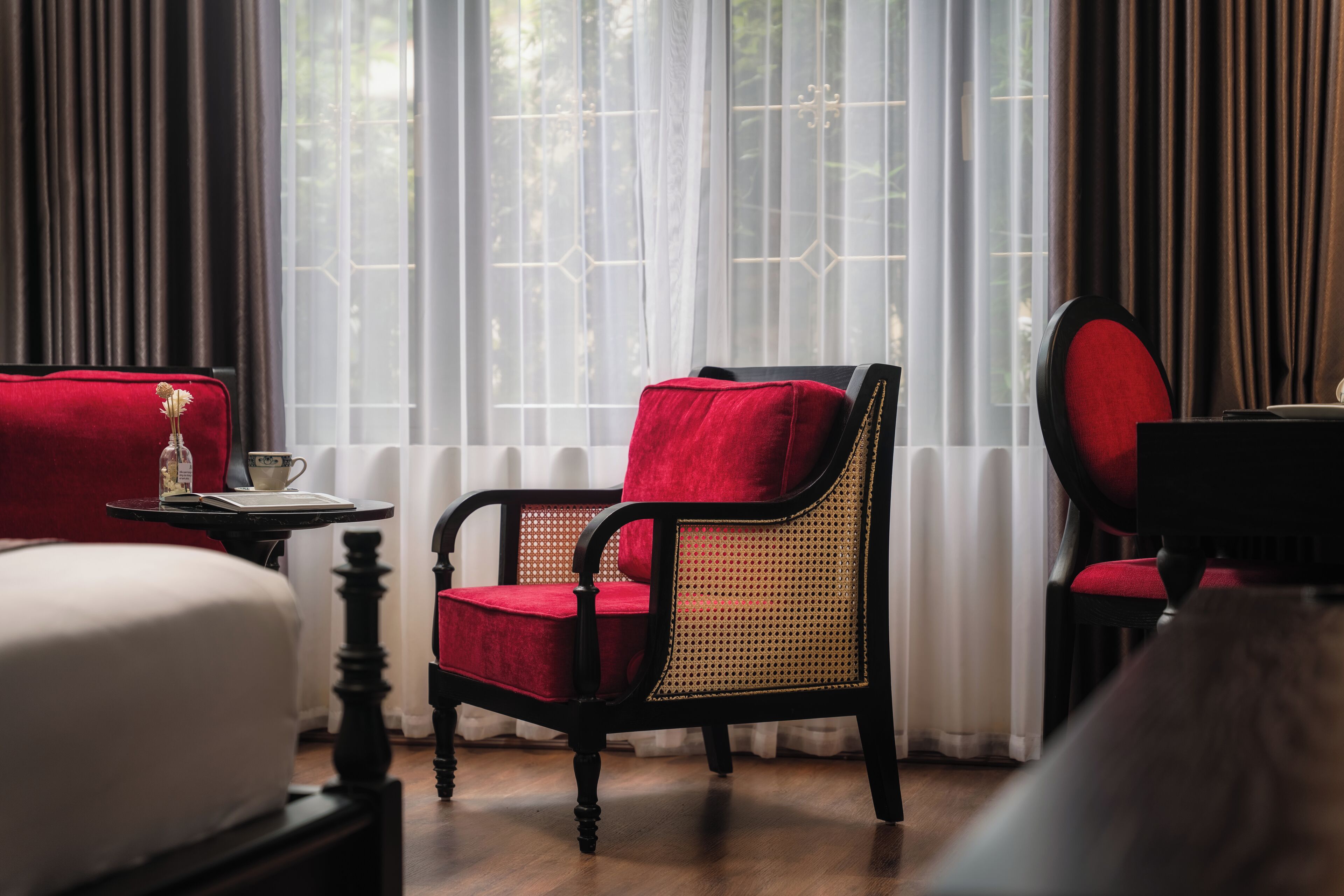 A hotel room with a red upholstered chair, a wooden table with a teapot and book, and a view through sheer curtains.
