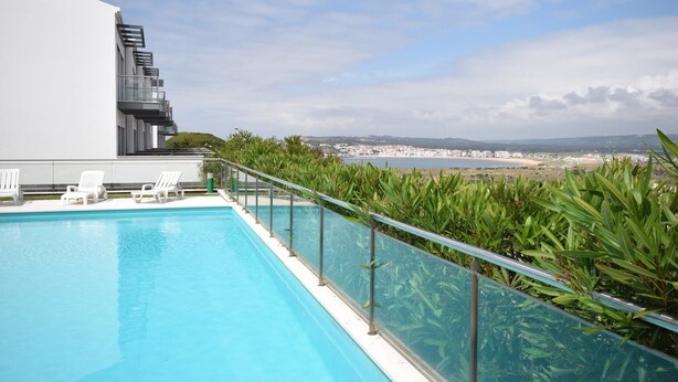 Ein Pool auf dem Dach mit weißen Liegestühlen, einer Glasuhr und Blick auf eine Küstenstadt.