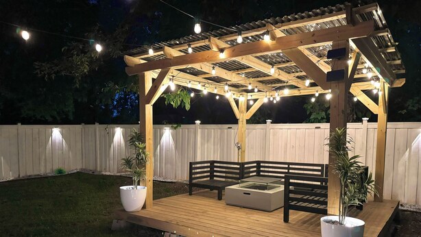 Eine Holzterrasse mit einer Pergola, Sitzgelegenheiten im Freien, Topfpflanzen und Lichterketten.