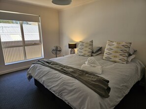 A neatly made bed with a blanket, pillows, and a rolled towel. There is a standing fan and a table lamp on a bedside table. - Bassett House Willunga - An entire spacious home on a tree lined street.  (Willunga)