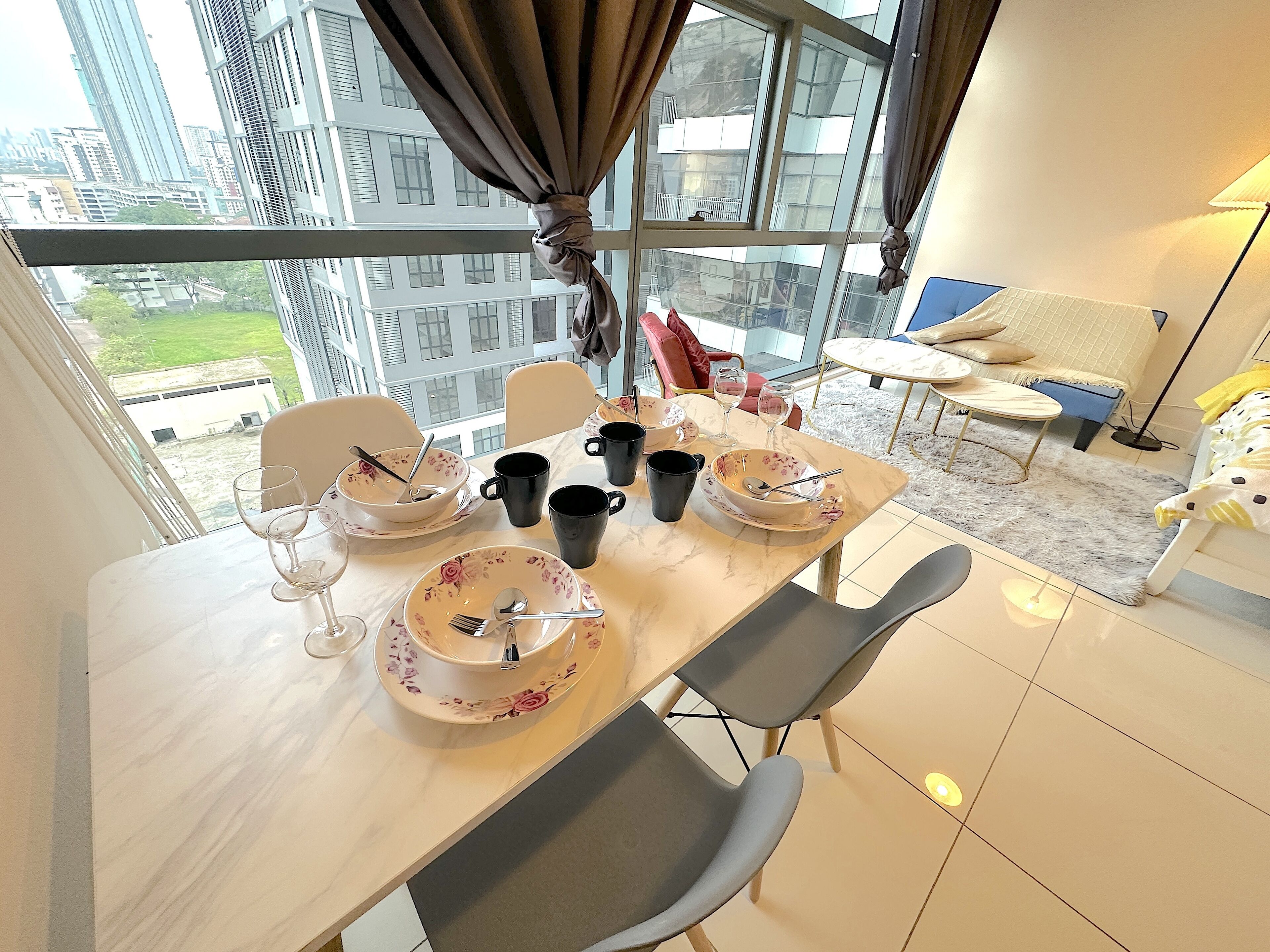 Un coin repas avec une table dressée pour deux personnes, un canapé et une vue sur le paysage urbain à travers de grandes fenêtres.
