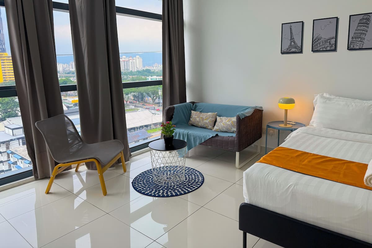 A hotel room with a bed, a sofa, a chair, a small table with a lamp, and a view of the city through large windows.