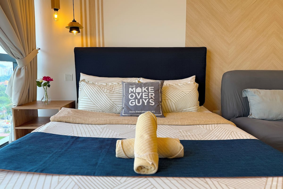 A neatly made bed with a pillow that reads "THE MAKE OVER GUYS" and a rolled towel on the bed.