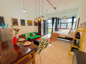 A modern living room with a sofa, dining table, and a bed. There are artworks on the walls, a window with a view of buildings, and a yellow shelf with items. - 3 Towers Jalan Ampang By Serenity Homes (Kuala Lumpur)