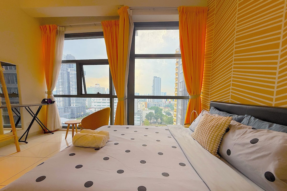 A hotel room with a large bed, a chair, a small table, and a view of the cityscape through large windows.