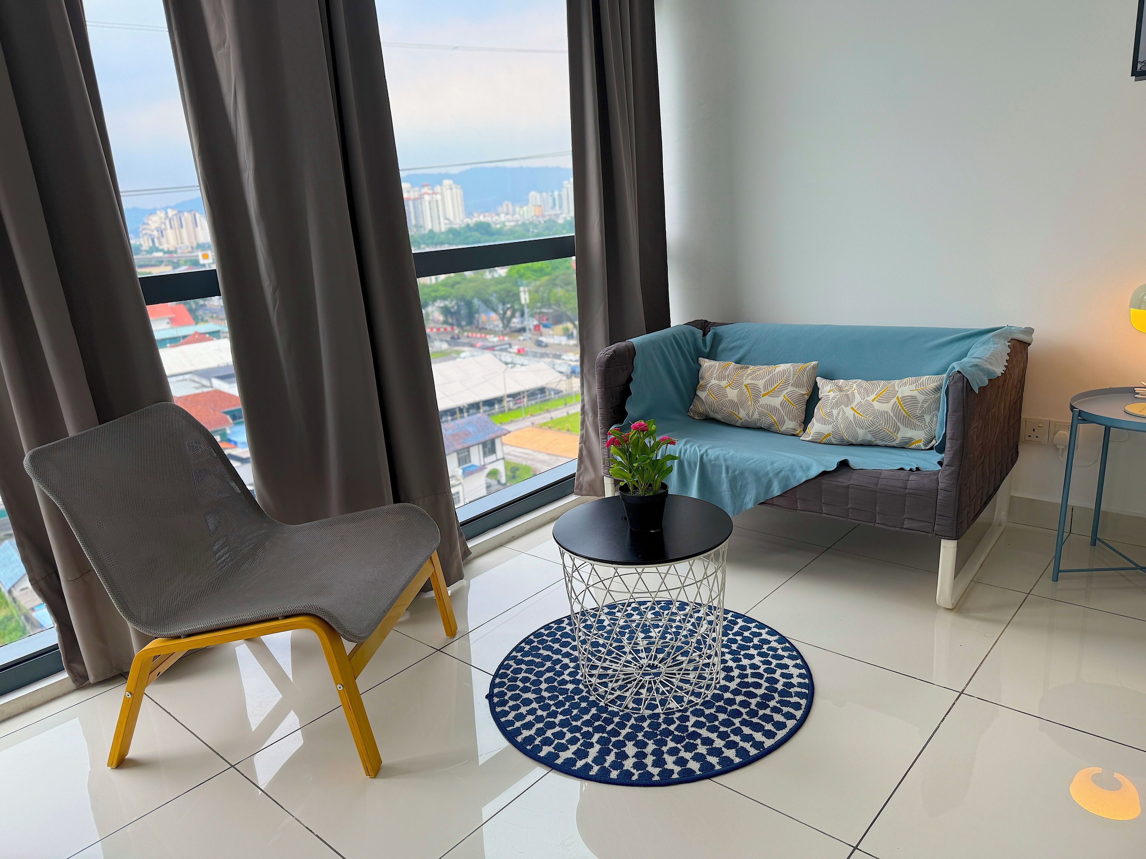 A modern living room with a sofa, a chair, a small table with a plant, and a view of a cityscape through large windows.