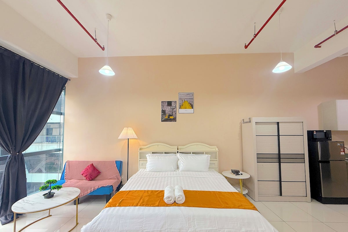 A hotel room with a bed, a small table, a chair, a lamp, a refrigerator, and a window with curtains.