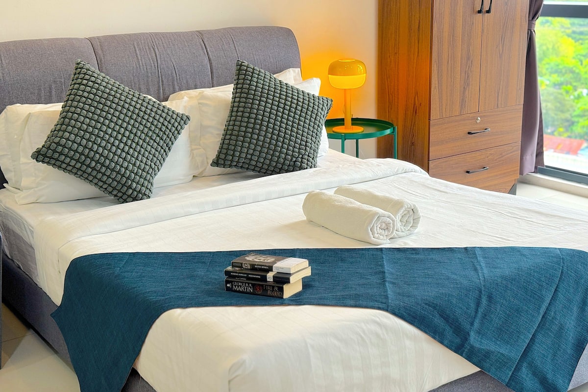 A neatly made bed with white linens, a teal throw blanket, and two rolled towels. There are two pillows with a textured pattern, a stack of books, and a wooden wardrobe in the background.