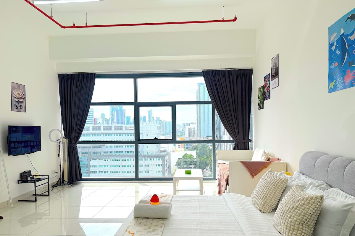 A modern hotel room with a large window, a bed with white bedding, a grey sofa, and a mounted TV.