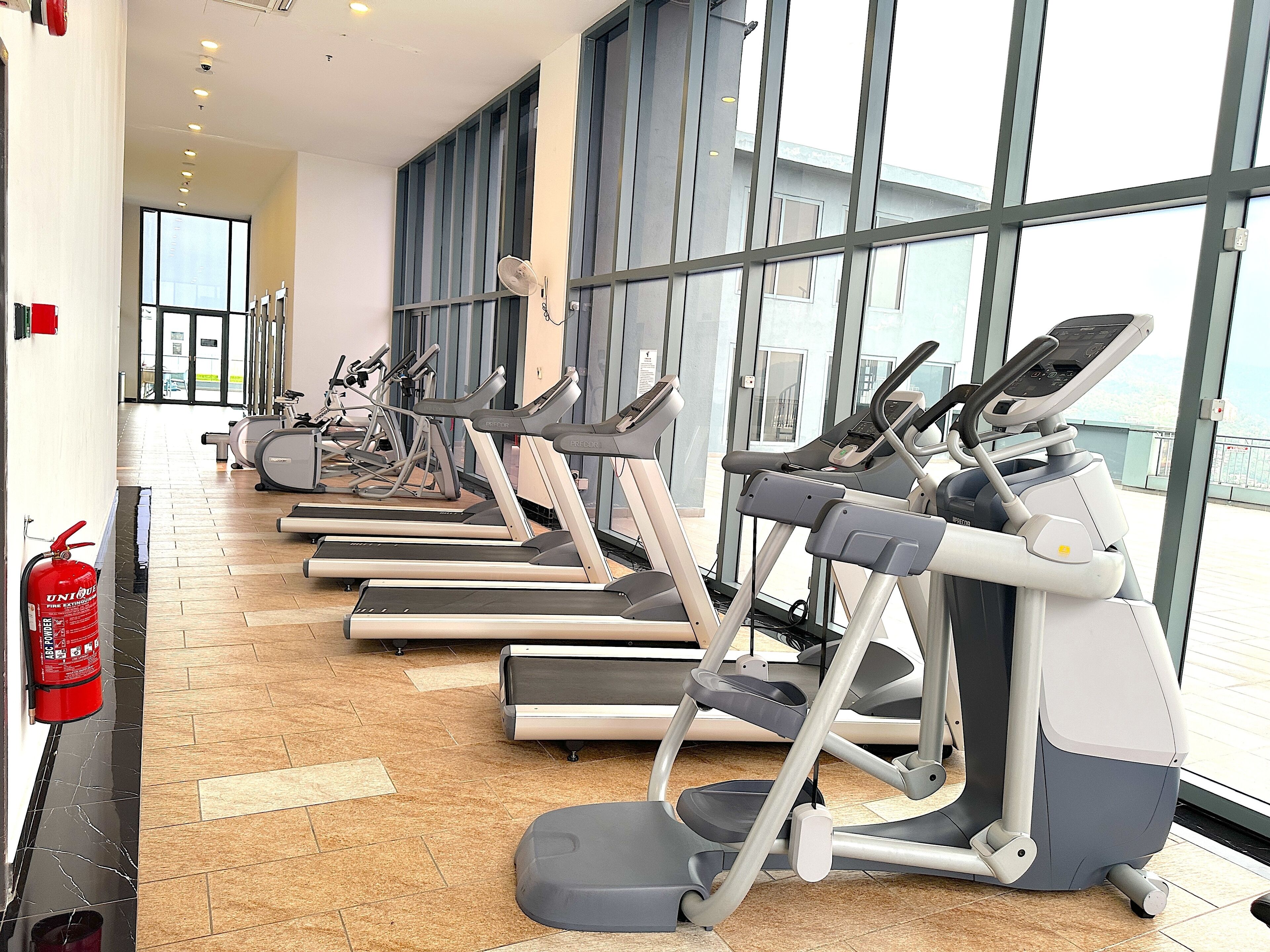 A gym with multiple treadmills aligned along a corridor with large windows and a fire extinguisher on the wall.
