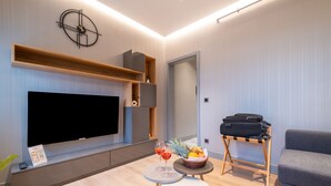 A modern living room with a large flat-screen TV, a wooden shelf, a small table with a basket of fruit, and a grey sofa. - The Platine Plus Suite (Istanbul)