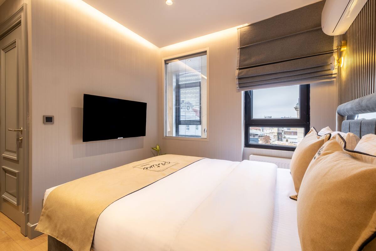 A modern hotel room with a large bed, a flat-screen TV mounted on the wall, a window with a view of buildings, and a ceiling-mounted air conditioner.