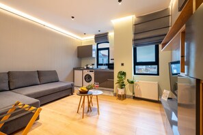 A modern living room with a grey sofa, a small round table with a fruit bowl and a glass, a washing machine, and a view of the city through large windows. - The Platine Plus Suite (Istanbul)